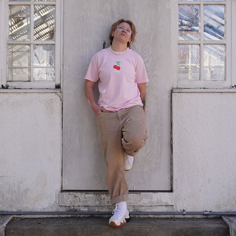 Cherry Fruity Shirt Outside Against Wall Abandoned House Light Pink T-Shirt Subtle Gay Pride Queer Fashion LGBTQ Clothing Inclusive Apparel